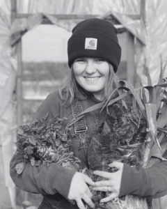 Holly, a Head Chef at Feast and Flow, smiling while holding freshly harvested leafy greens, wearing a beanie and standing in a greenhouse.